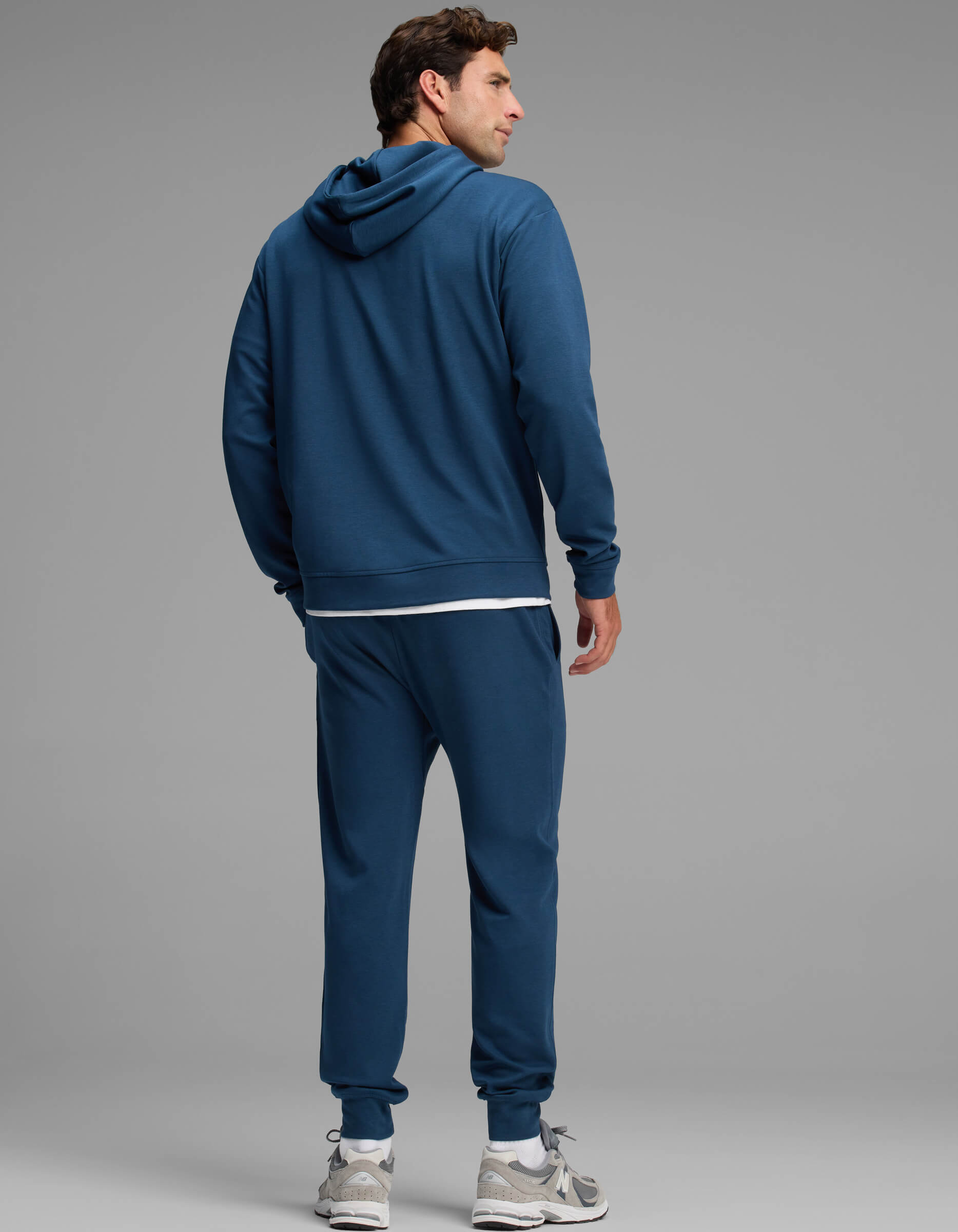 A man stands facing away, wearing the Cozy Earth Men's Versa Hoodie & Jogger Set in blue, paired with white sneakers against a plain gray background.