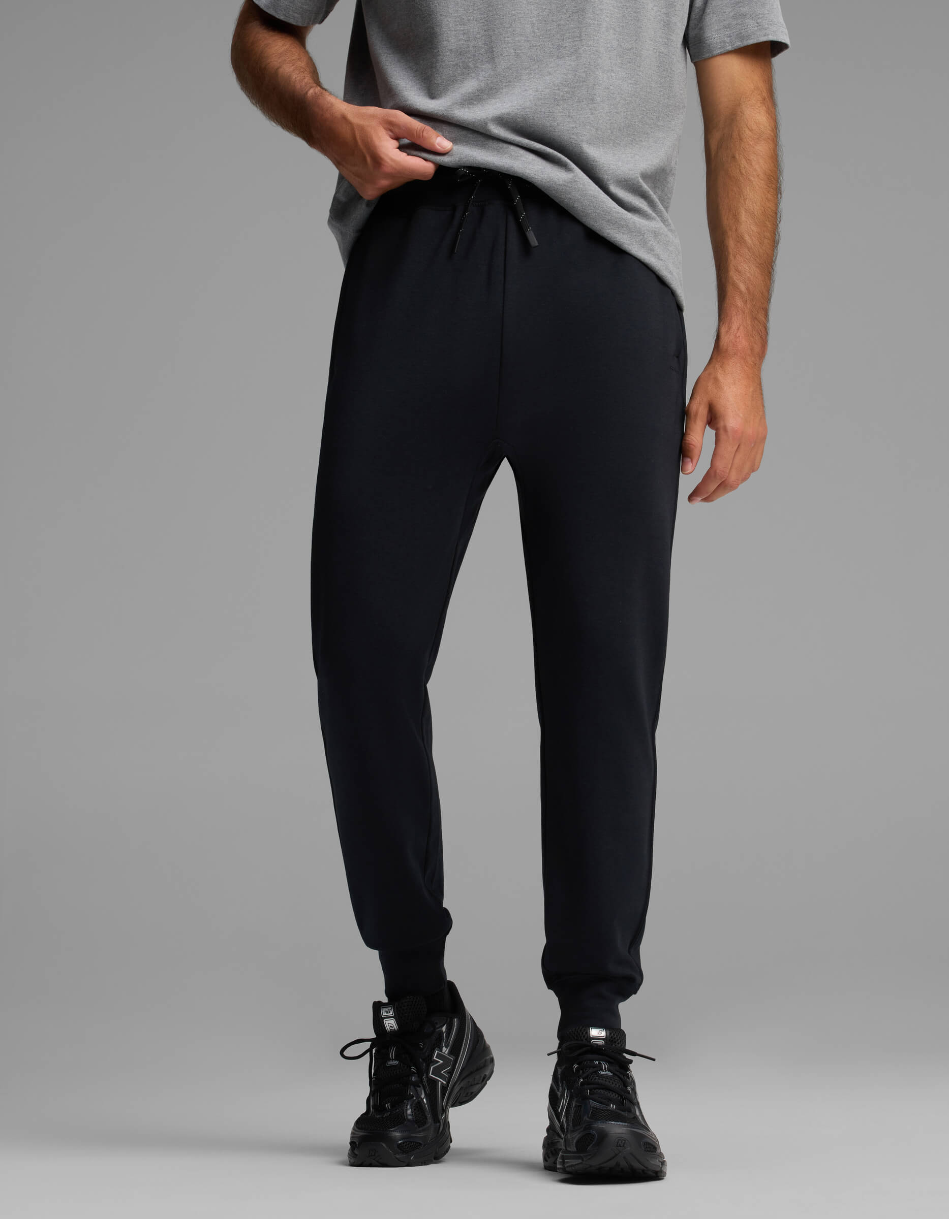 A person wearing Cozy Earth's Men's Versa Jogger in black and a grey T-shirt stands against a plain grey background, holding the hem of their shirt, with only their lower torso and legs visible.