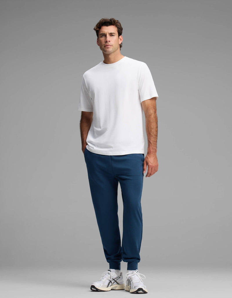 A man stands against a gray background wearing a plain white t-shirt, white sneakers, and Cozy Earth's Men's Versa Jogger in blue, with his left hand in his pocket.