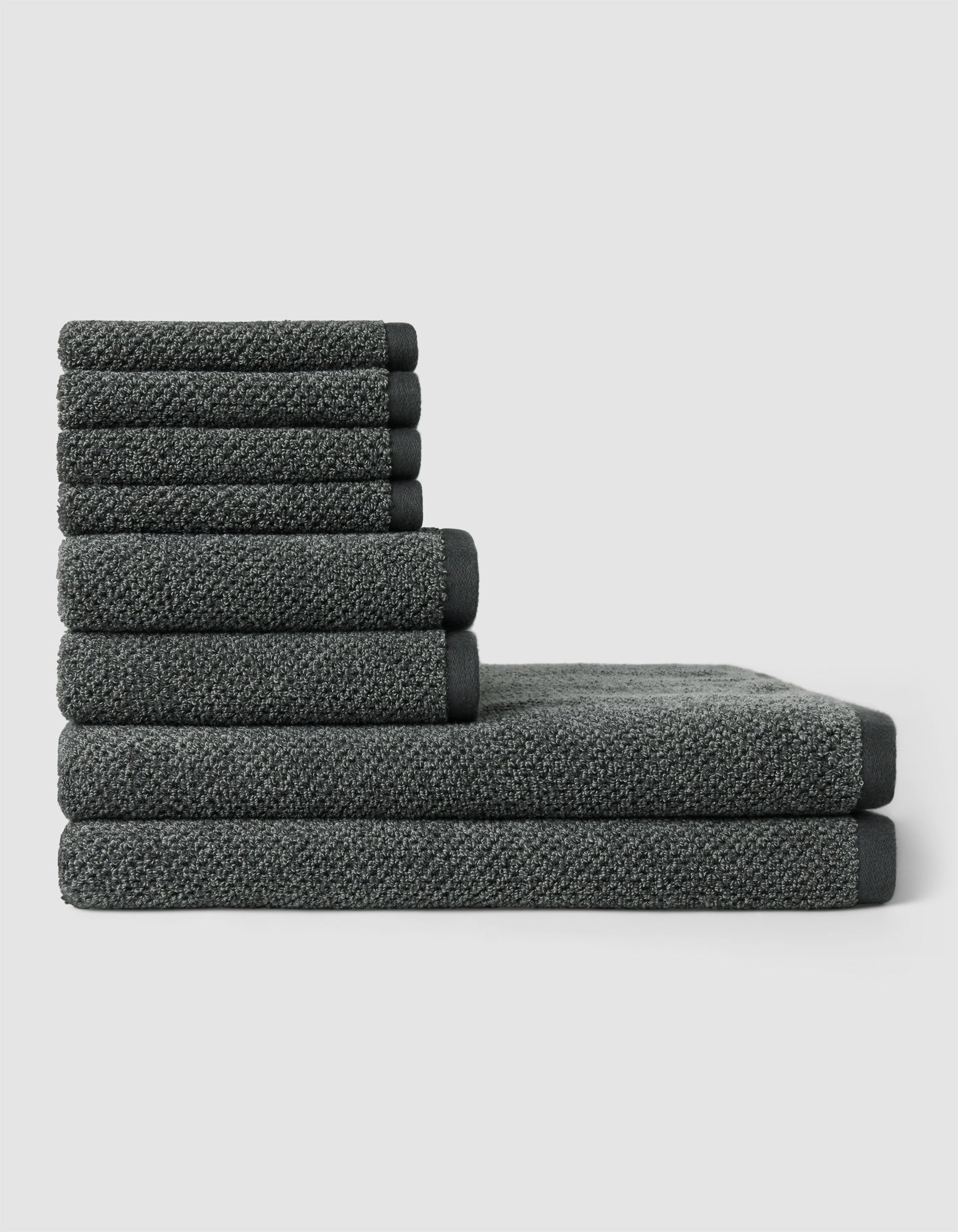 The Nantucket Bath Towel Set by Cozy Earth features dark gray towels in various sizes, neatly stacked in a step-like pattern against a plain light gray background.|Color:Graphite