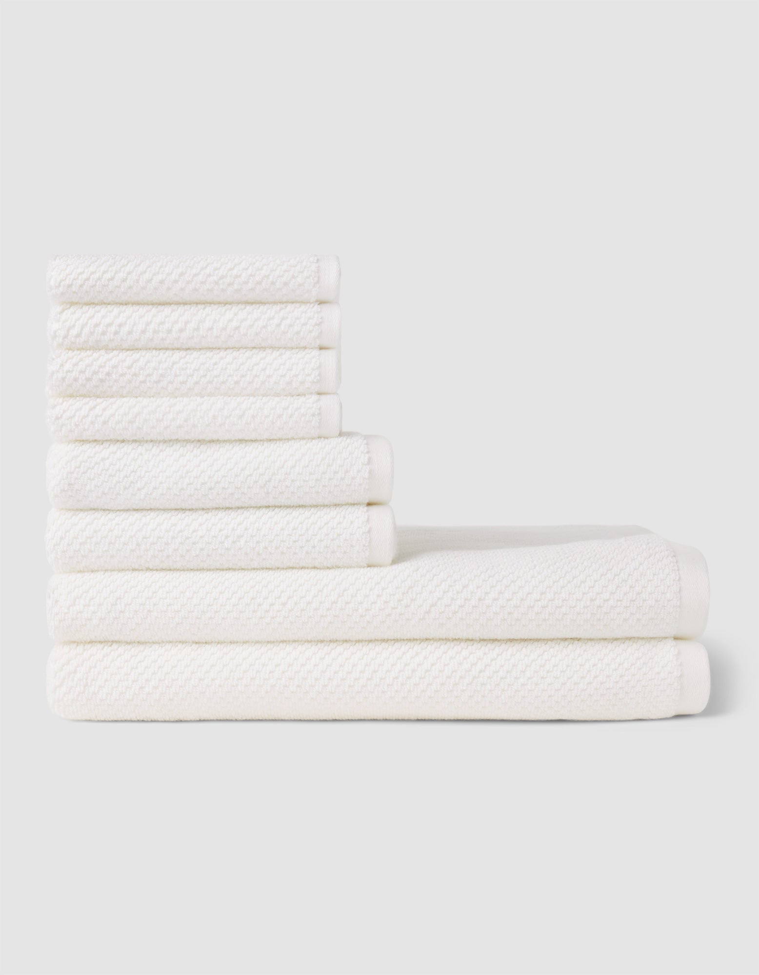 The Cozy Earth Nantucket Bath Towel Set features seven white textured towels—three large and four small—neatly stacked in two rows on a light gray background, showcasing their soft, plush feel.|Color:White