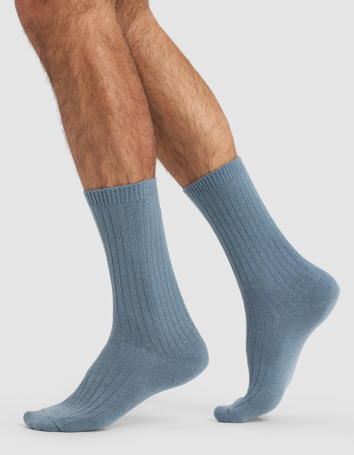 A person with hairy legs models Cozy Earth's The Plush Lounge Calf Sock 3-Pack in light blue, ribbed crew style, standing on a plain white background with one foot flat and the other slightly raised, toes pointed.