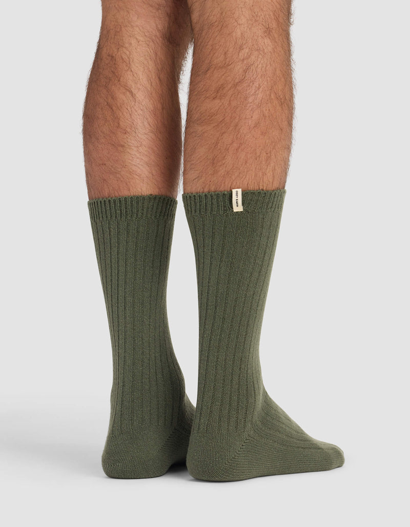 A person stands on a light surface wearing Cozy Earth's Men's Plush Lounge Calf Sock in olive green, featuring ribbing and a small beige tag near the top. Only their lower legs and feet are visible.