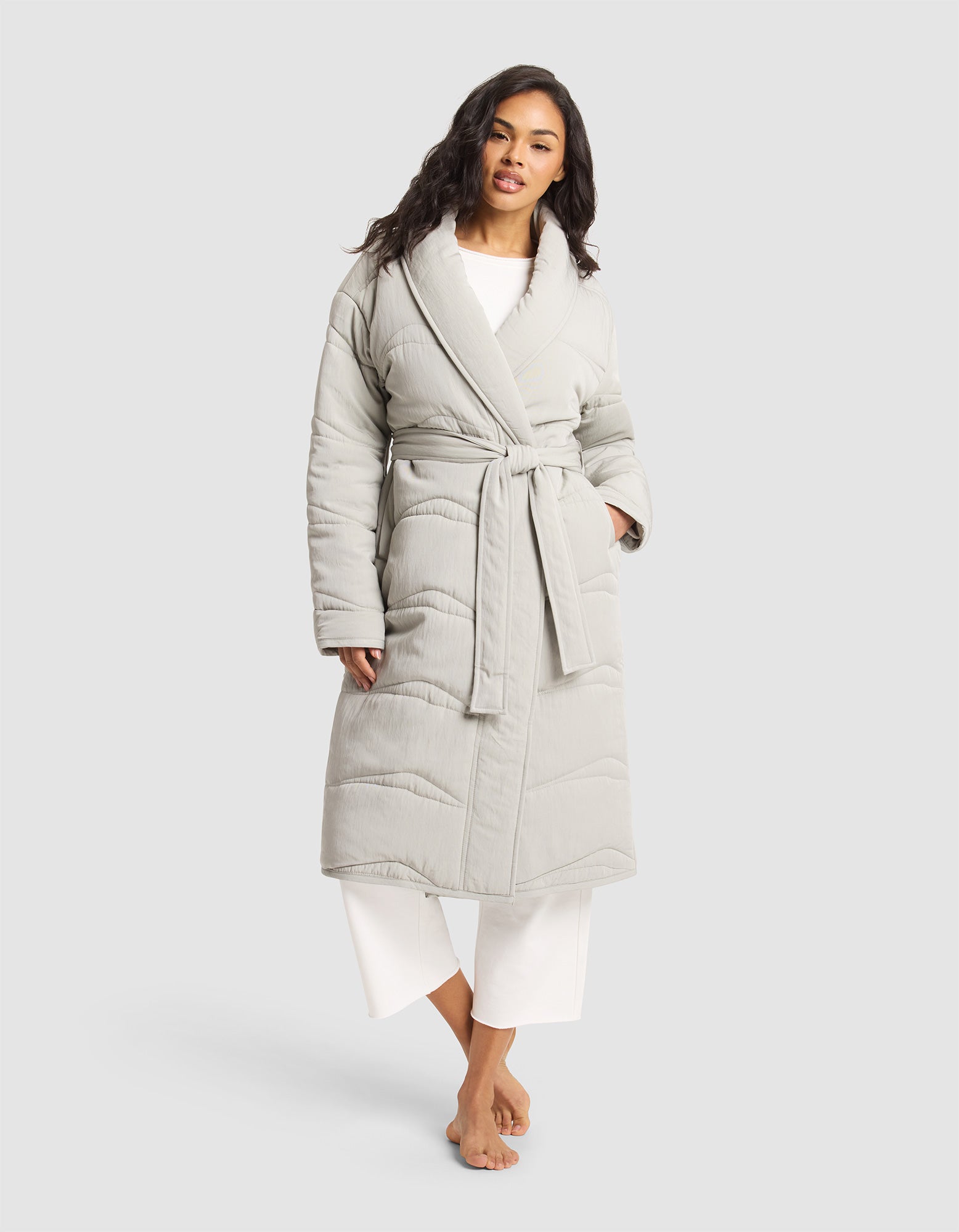 A woman stands barefoot against a plain light background, wearing Cozy Earth's Women's Quilted House Coat in light gray with a tied belt, layered over white pajama pants. 