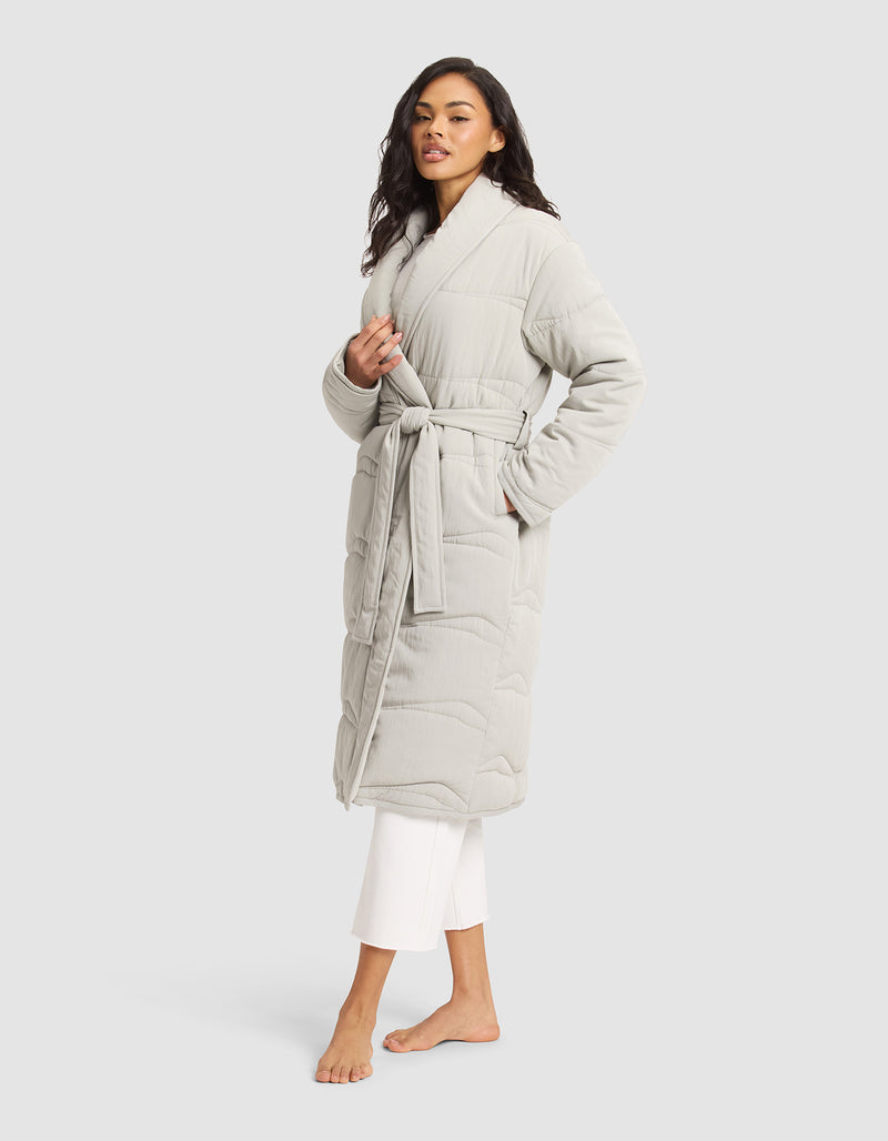 A woman stands barefoot against a plain light background wearing Cozy Earth's Women's Quilted House Coat, a long, light gray robe with a belt tie, styled over white pants.
