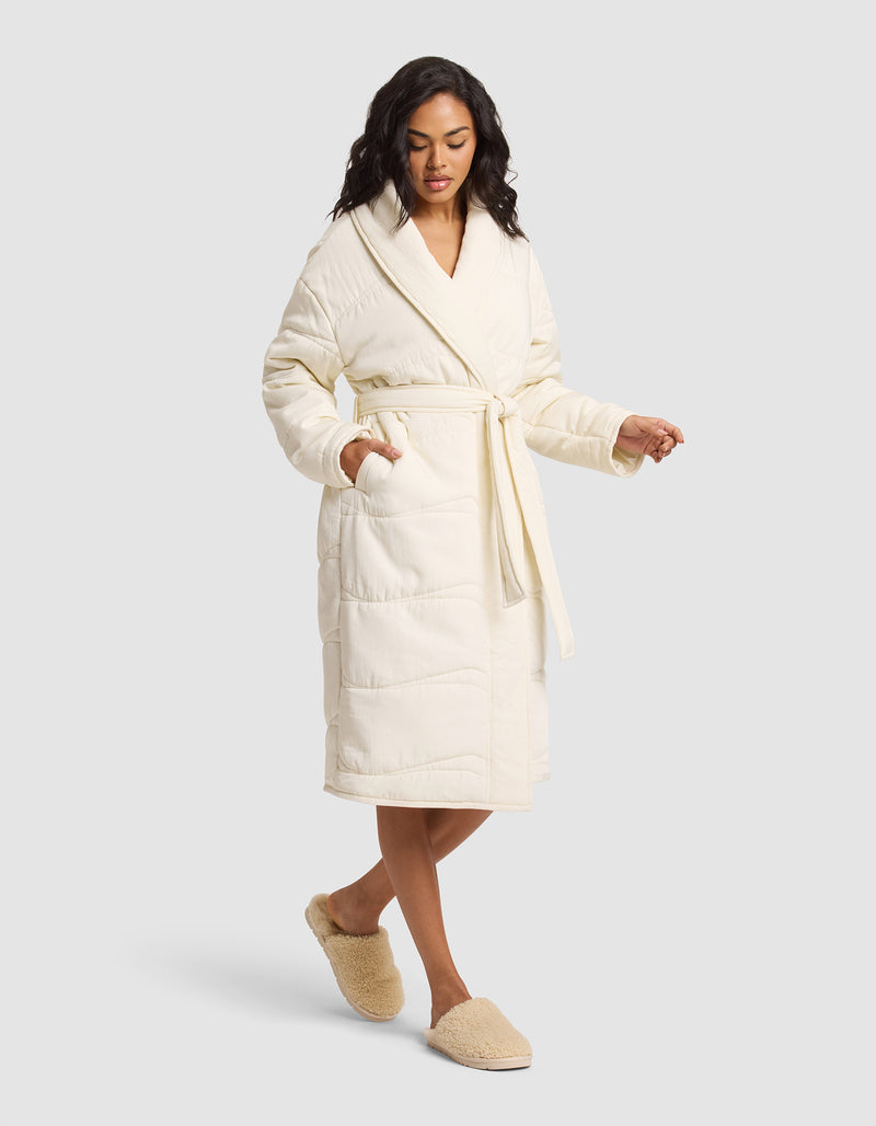 A woman models the Cozy Earth Women's Quilted House Coat, a long cream robe tied at the waist, paired with beige fuzzy slippers, standing against a plain light gray background.