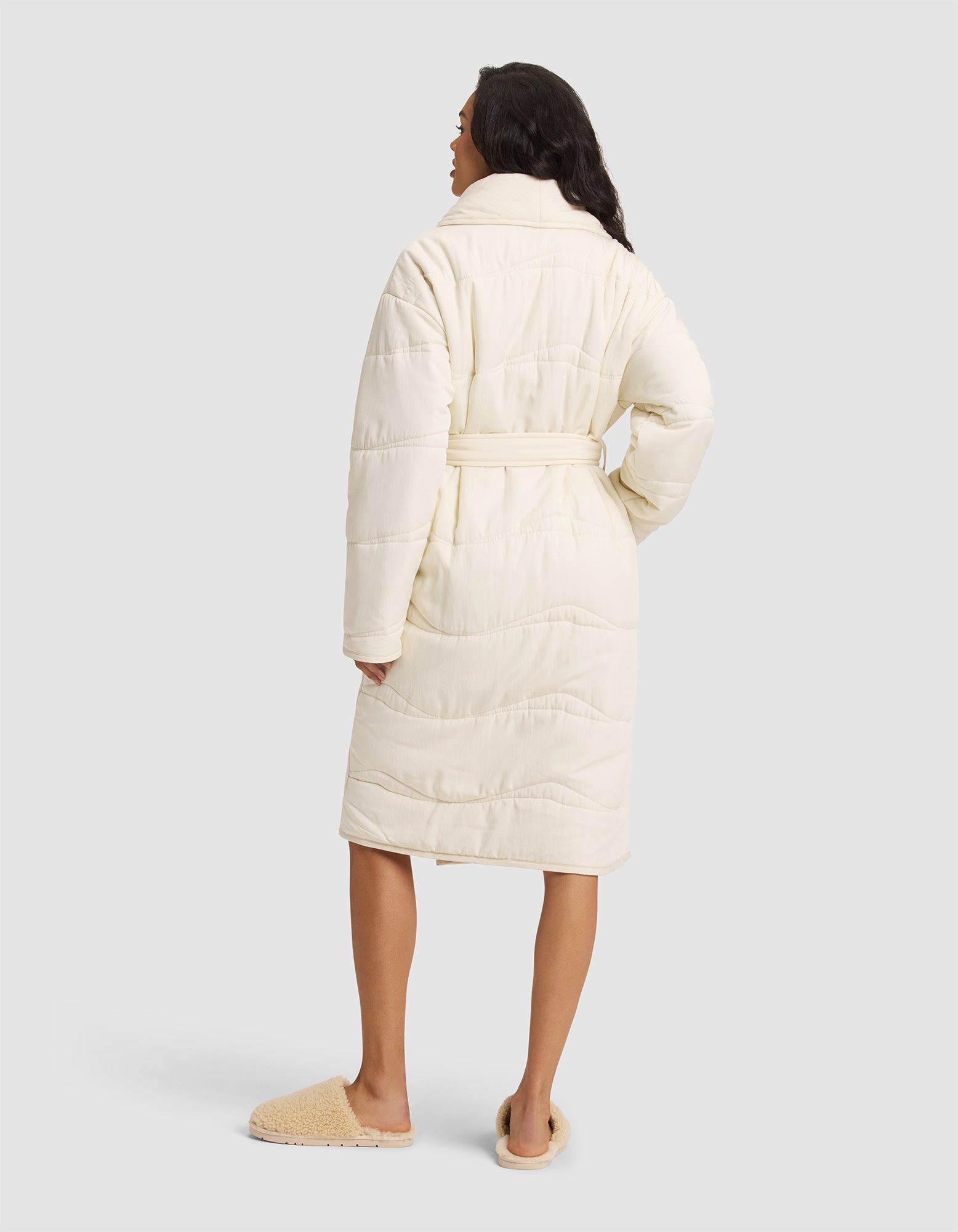A woman faces away, wearing the Cozy Earth Women's Quilted House Coat in cream with a belt and tan slippers, set against a plain light gray background. 