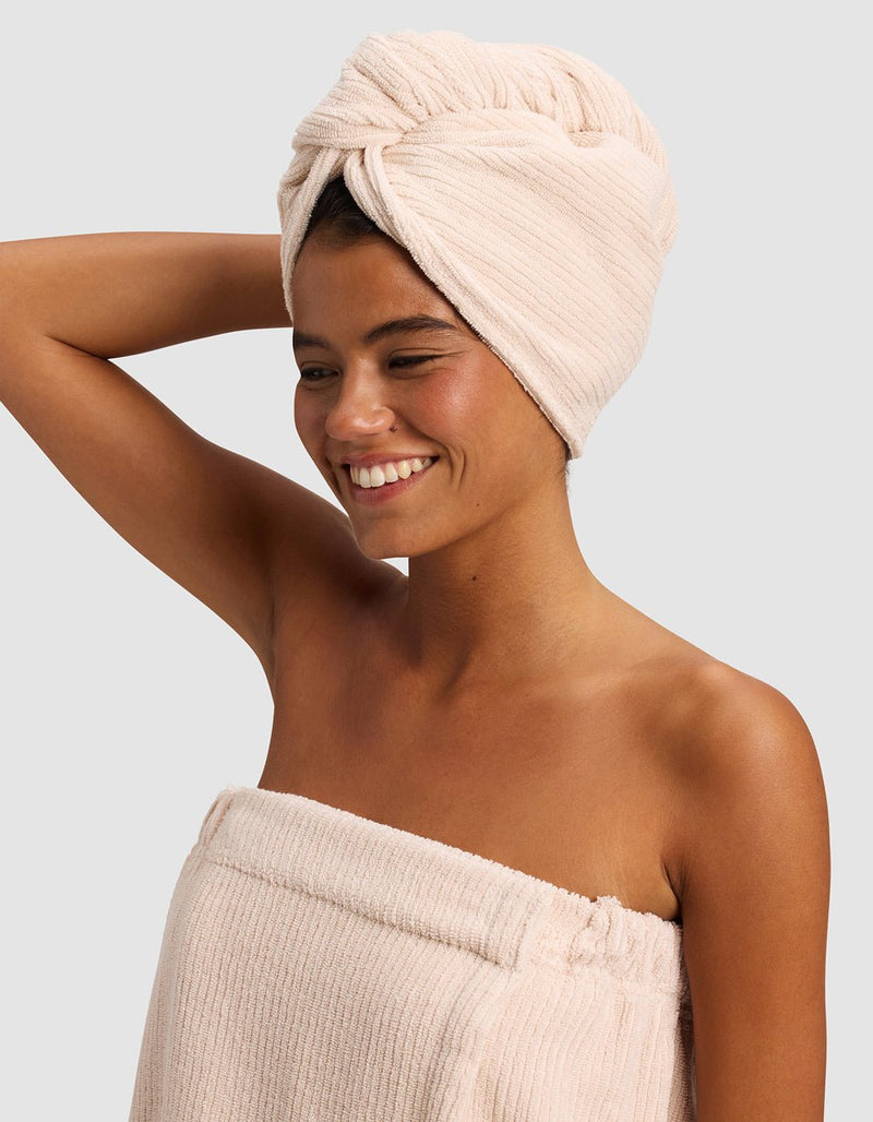 A smiling woman stands against a light gray background, wearing a beige towel wrap and the Cozy Earth Ribbed Terry Hair Towel in matching beige, with one arm raised and bent behind her head.