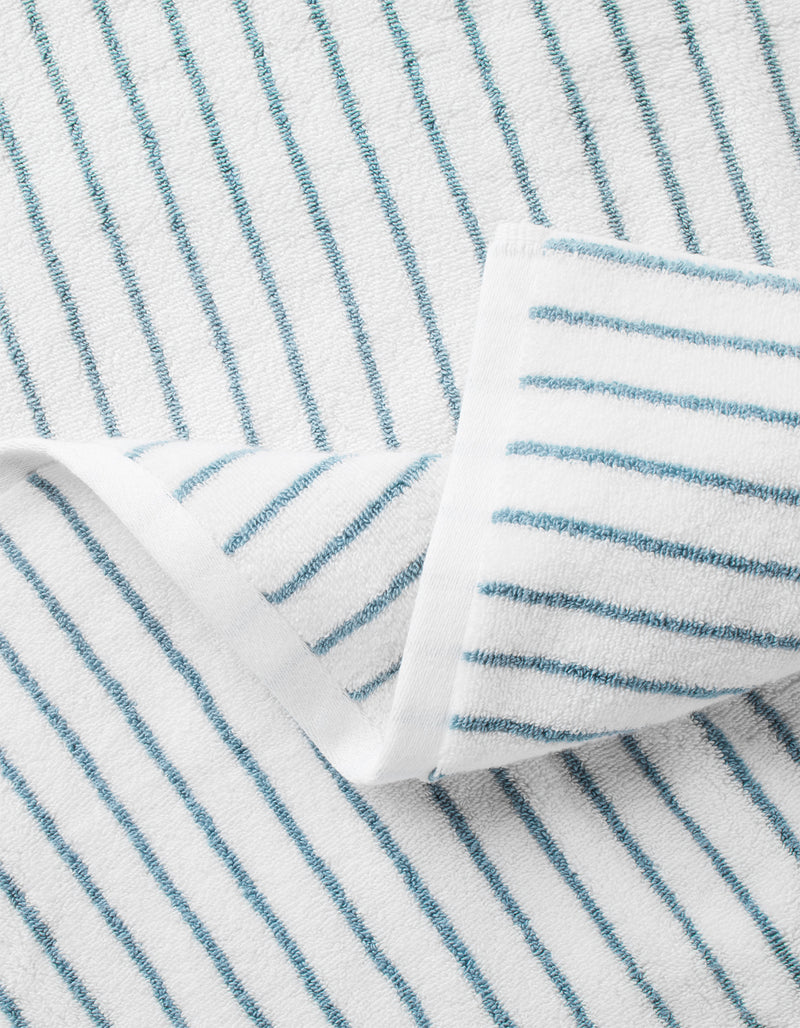 A close-up of Cozy Earth's Striped Luxe Bath Sheets in white with thin blue diagonal stripes, featuring a folded corner to highlight the textured detail.