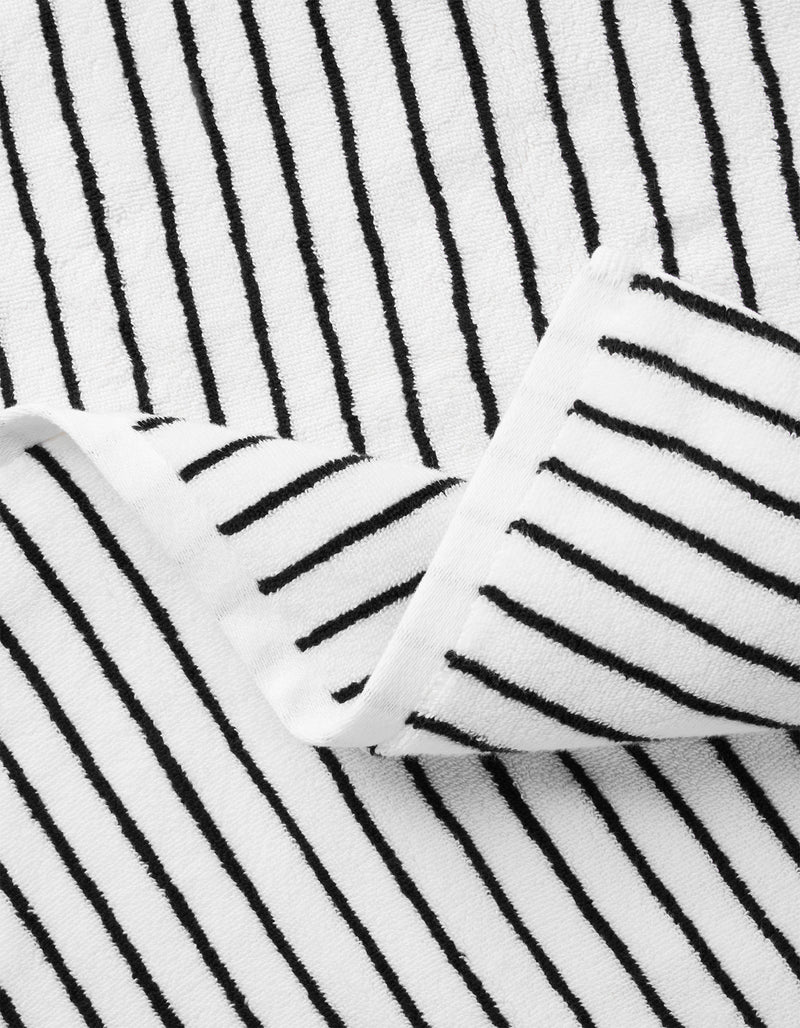 A close-up of Cozy Earth's Striped Luxe Bath Sheets shows white fabric with thin, evenly spaced black stripes. The slight folds in the bath sheets create overlapping sections and soft shadows.