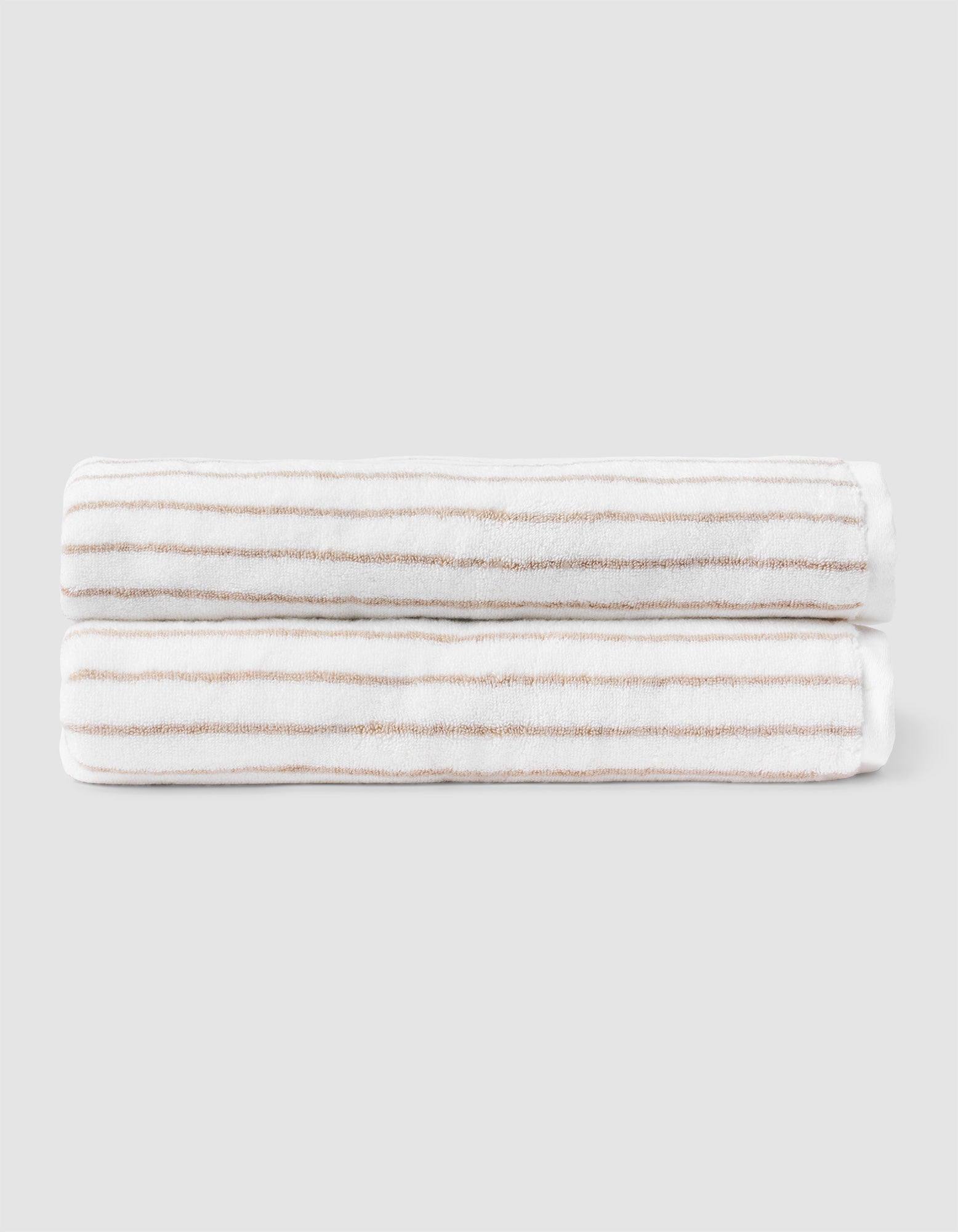 Two Striped Luxe Bath Sheets by Cozy Earth, featuring white fabric with thin, evenly spaced tan stripes, are neatly folded and stacked against a plain light gray background.  |Color:Sand Stripe