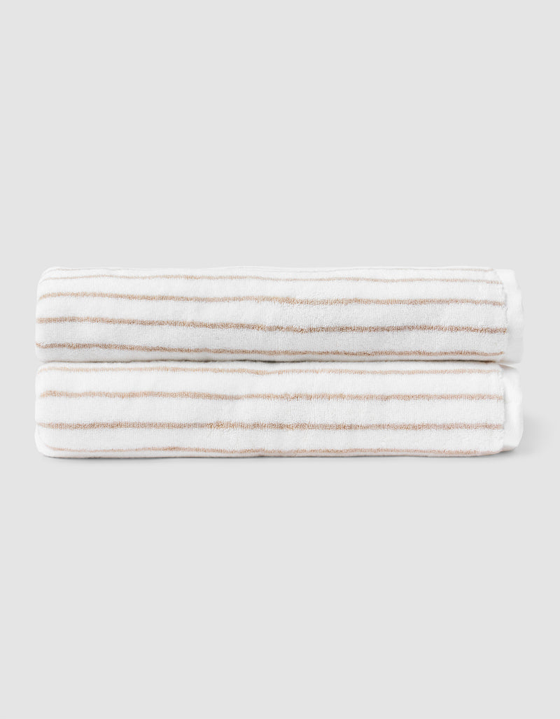 Two Striped Luxe Bath Sheets by Cozy Earth, featuring white fabric with thin, evenly spaced tan stripes, are neatly folded and stacked against a plain light gray background.
