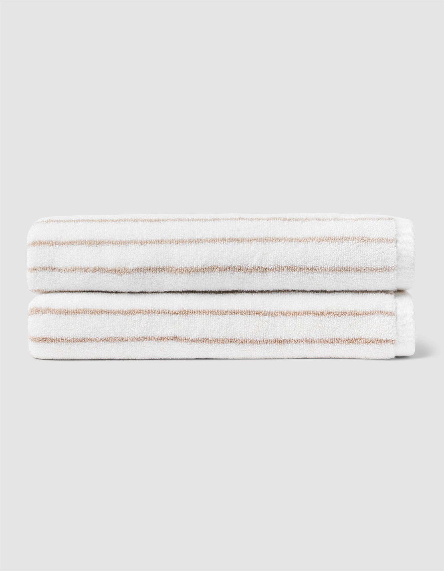 Two Striped Luxe Bath Towels by Cozy Earth, featuring thin light brown stripes on white, are stacked neatly against a plain light gray background. |Color:Sand Stripe