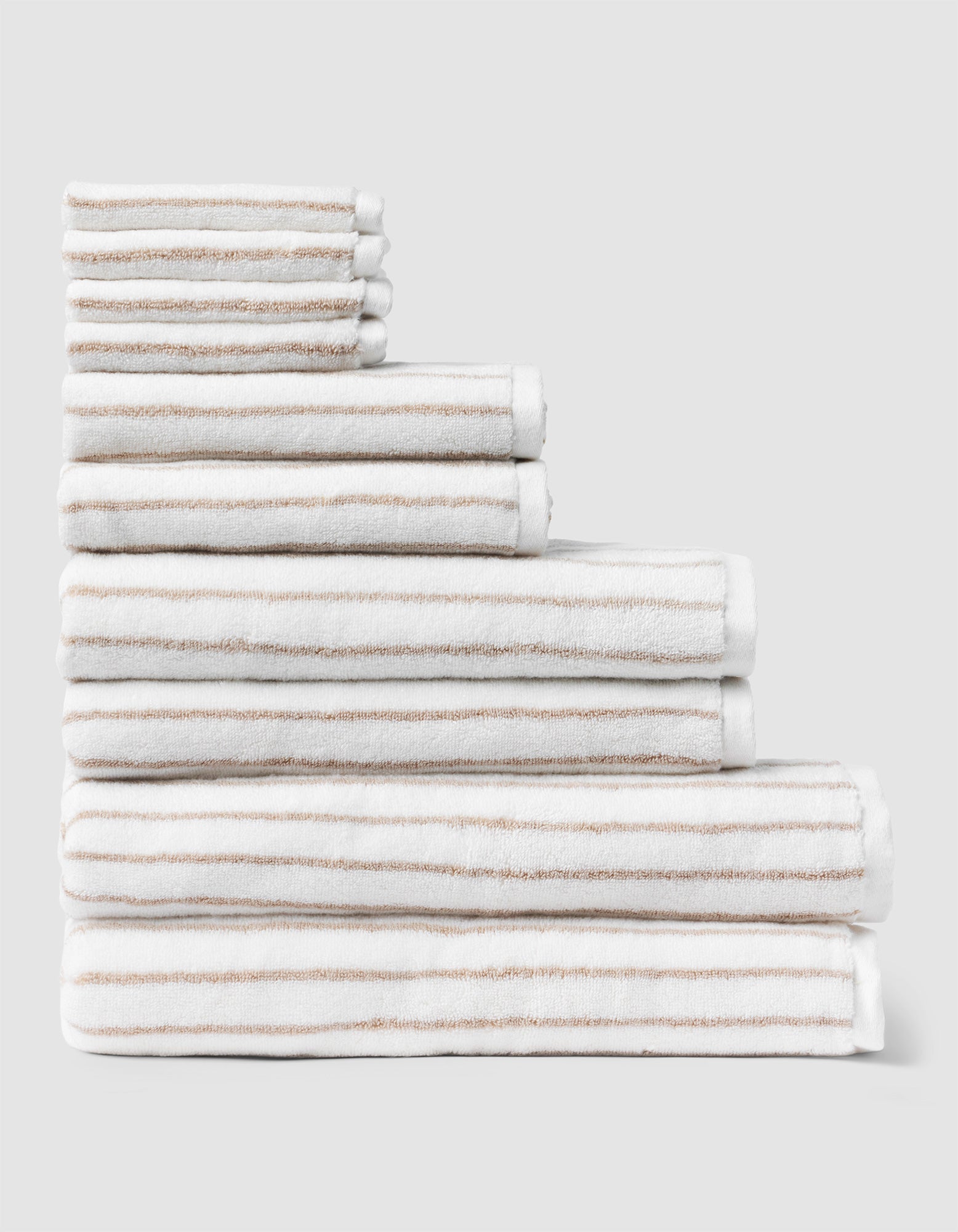 The Complete Striped Luxe Bath Bundle by Cozy Earth features neatly stacked white towels with thin beige stripes, arranged in descending size order against a plain light gray background. |Color:Sand Stripe