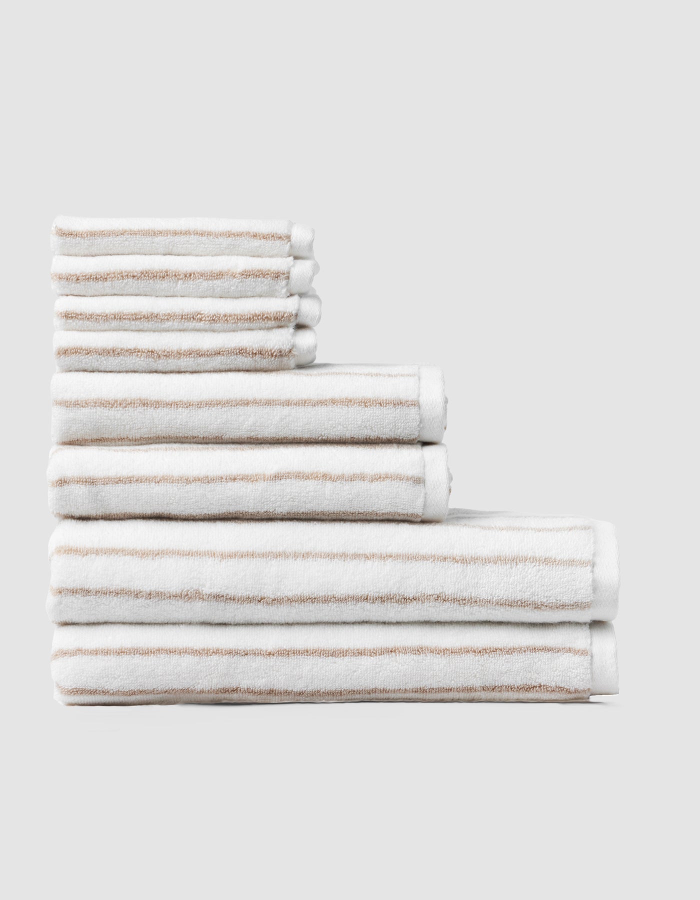 The Cozy Earth Striped Luxe Bath Towel Set features neatly folded white towels with beige stripes in three sizes, displayed against a plain light gray background. |Color:Sand Stripe