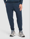 A person wears Cozy Earth's Men's StretchTech Quarter Zip & Jogger Set in navy, paired with light gray sneakers, standing with one hand in their pocket against a plain light background.A man stands against a plain background wearing Cozy Earth’s Men's StretchTech Jogger in black, paired with a black sweatshirt and gray slippers. He has one hand in his pocket and looks slightly to the side. |Color:Eclipse