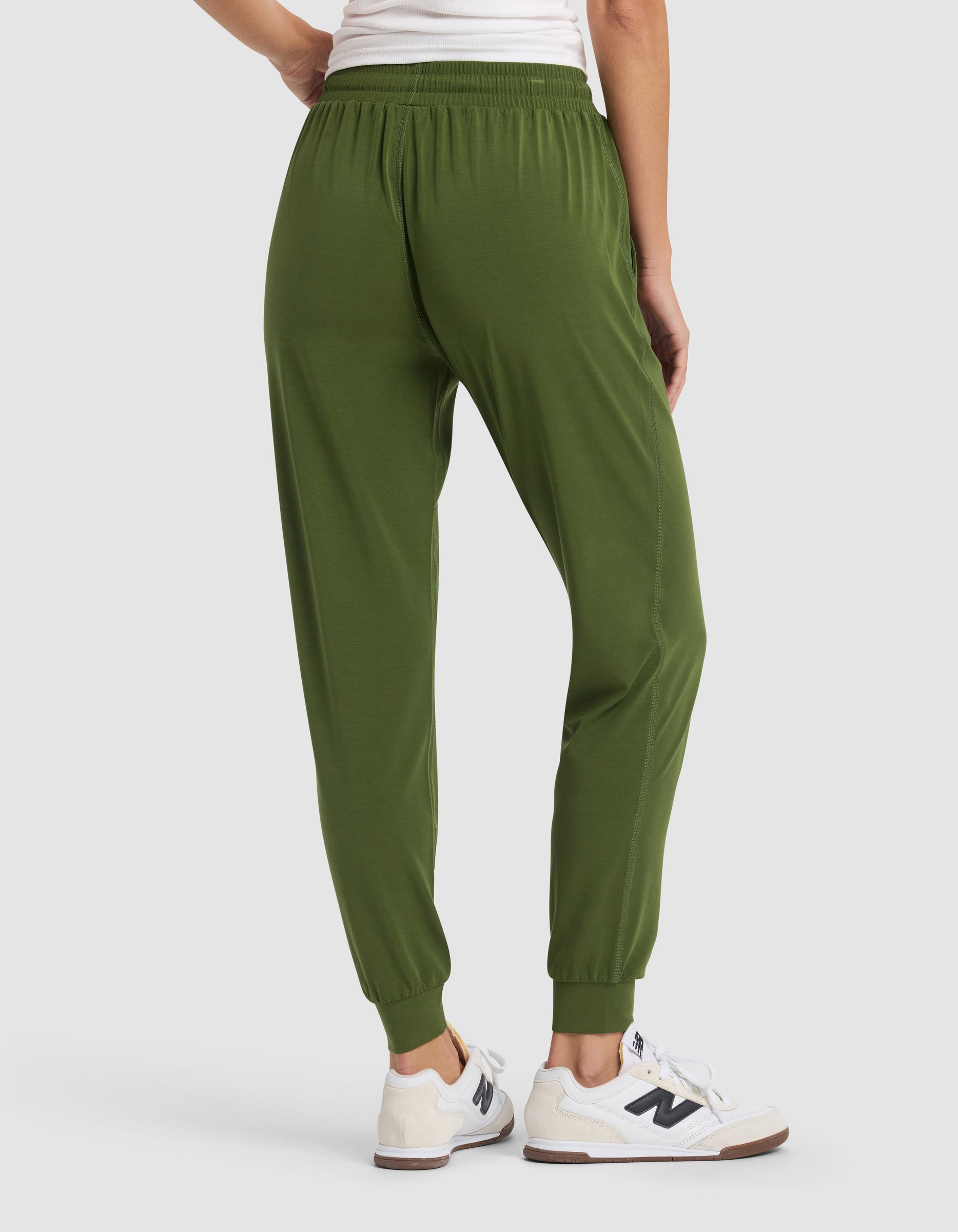 A woman is shown from the waist down wearing Cozy Earth Women's Studio Jogger pants in olive green, paired with white sneakers, standing on a light gray background.