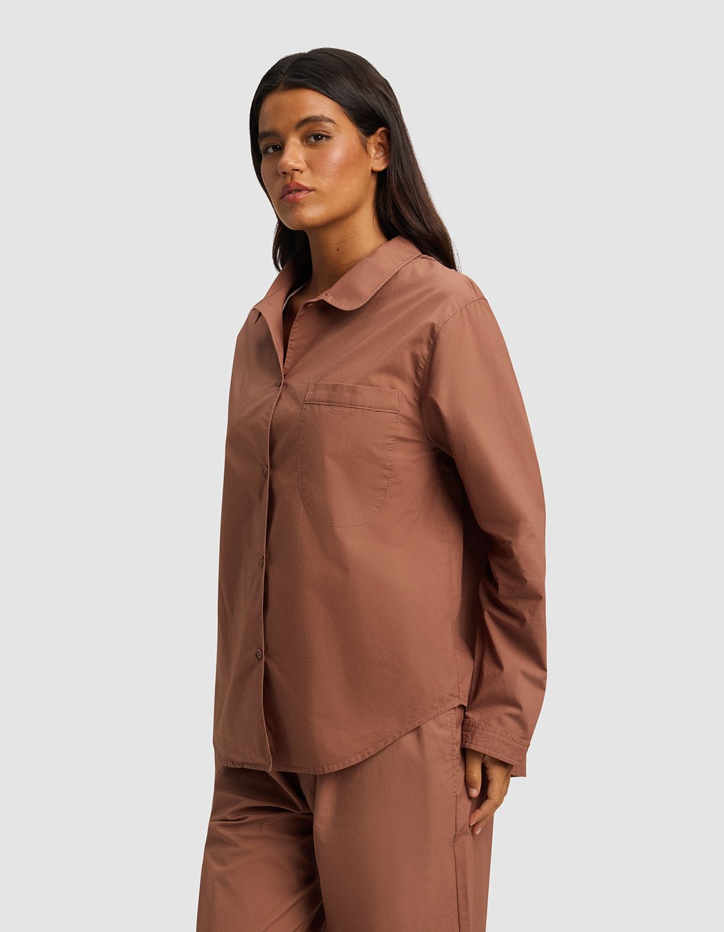 A woman with long dark hair confidently wears the Cozy Earth Women's Sutton Long Sleeve Pajama Top in dusty rose, paired with matching pants, standing against a plain light gray background.