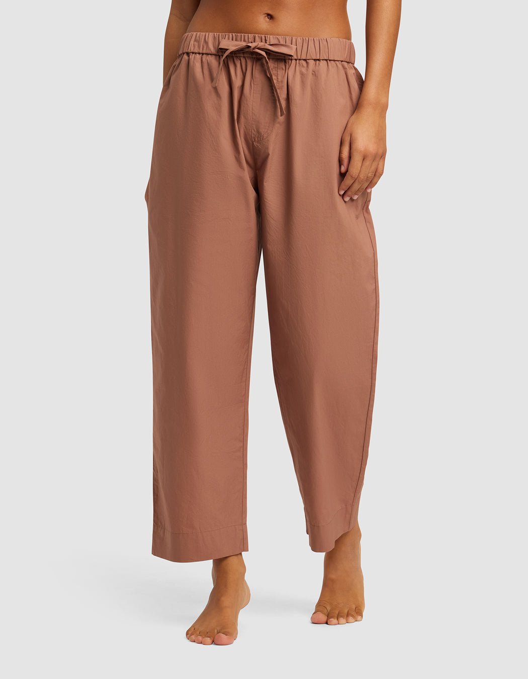 A woman models Cozy Earth's Women's Sutton Cropped Pajama Pant—loose, light brown, ankle-length with an elastic waistband and drawstring—while standing barefoot against a plain background.