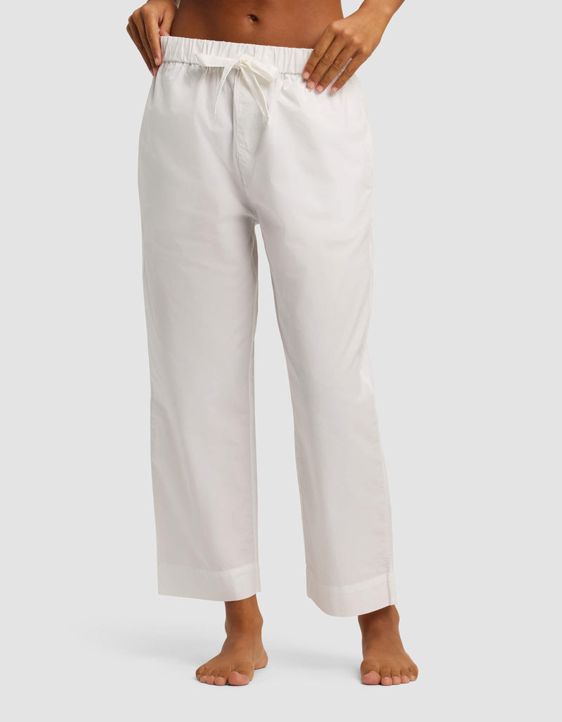 A woman stands barefoot against a plain light background, wearing Cozy Earth's Women's Sutton Short Sleeve & Pant Pajama Set with loose-fitting white drawstring pants cropped at the ankles. The upper body is partially out of frame.