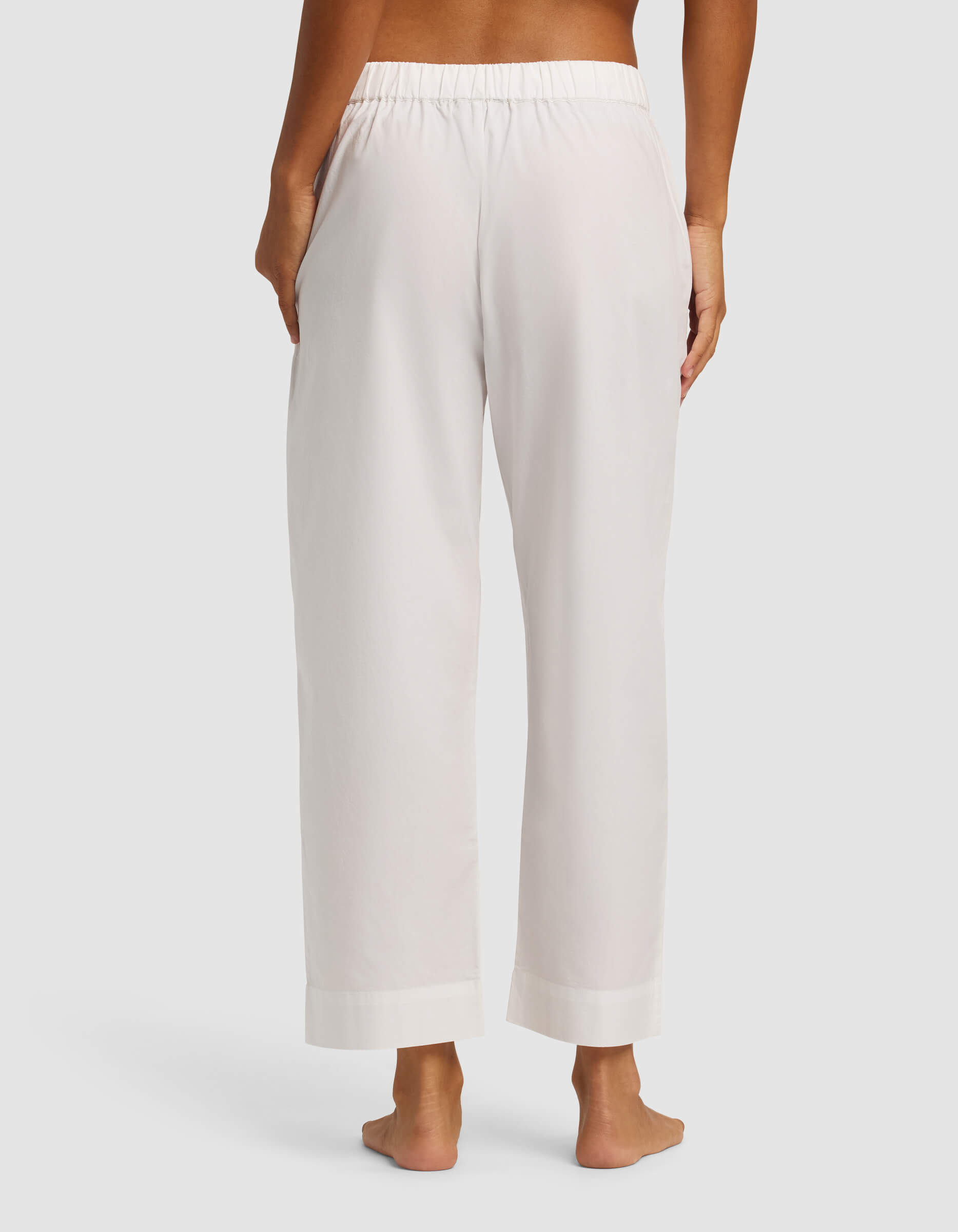 A woman stands barefoot, facing away from the camera, wearing Cozy Earth's Women's Sutton Cropped Pajama Pant—loose-fitting, light-colored with an elastic waistband and pockets—against a plain light background.