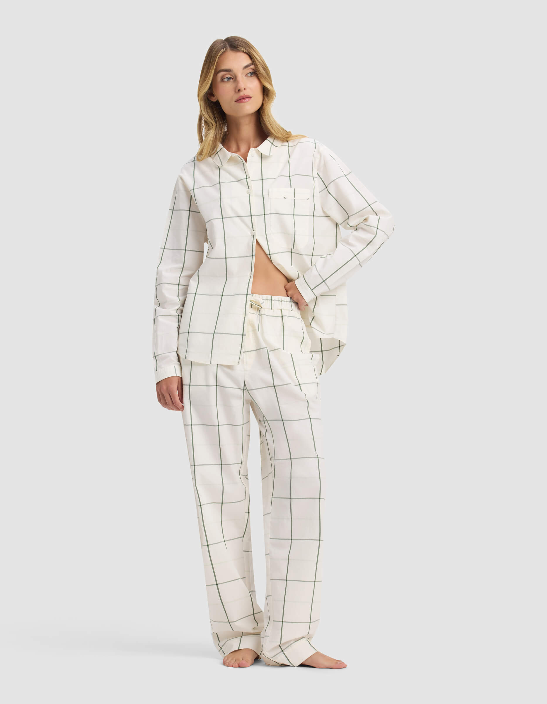A woman stands barefoot against a light gray background, wearing Cozy Earth's Women's Sutton Pajama Pant in white with a black grid pattern and a loose button-up shirt partly tucked in, revealing her midriff.