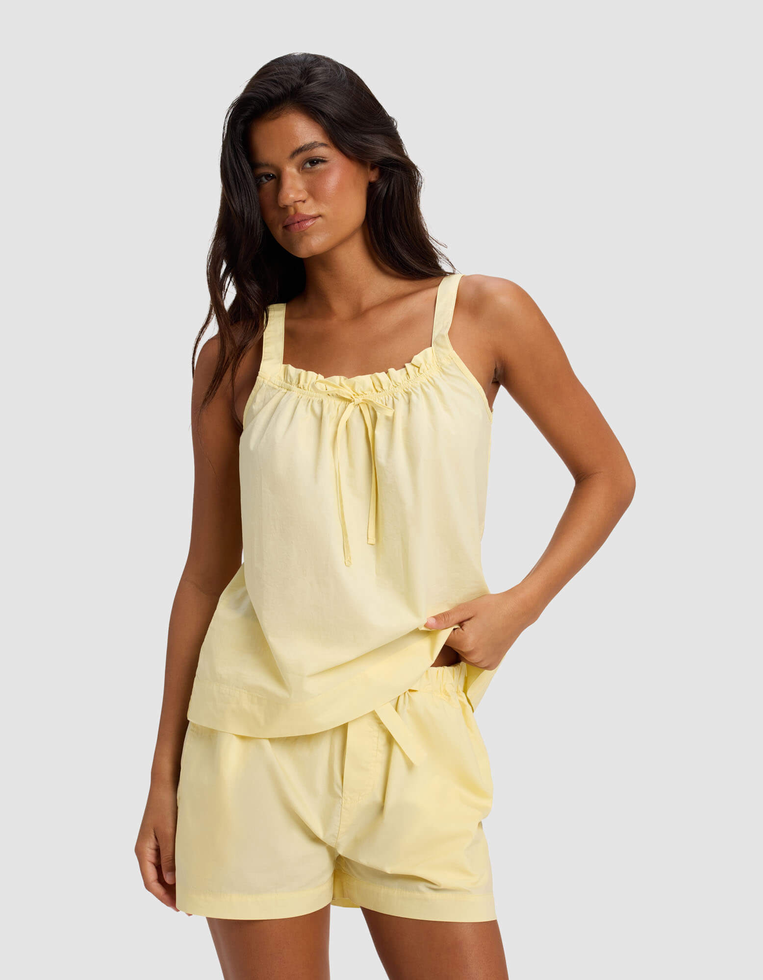 A woman wears Cozy Earth's Women's Sutton Pajama Tank in Meringue, featuring a tie neckline, paired with matching loose shorts, standing against a plain light gray background.|Color:Meringue