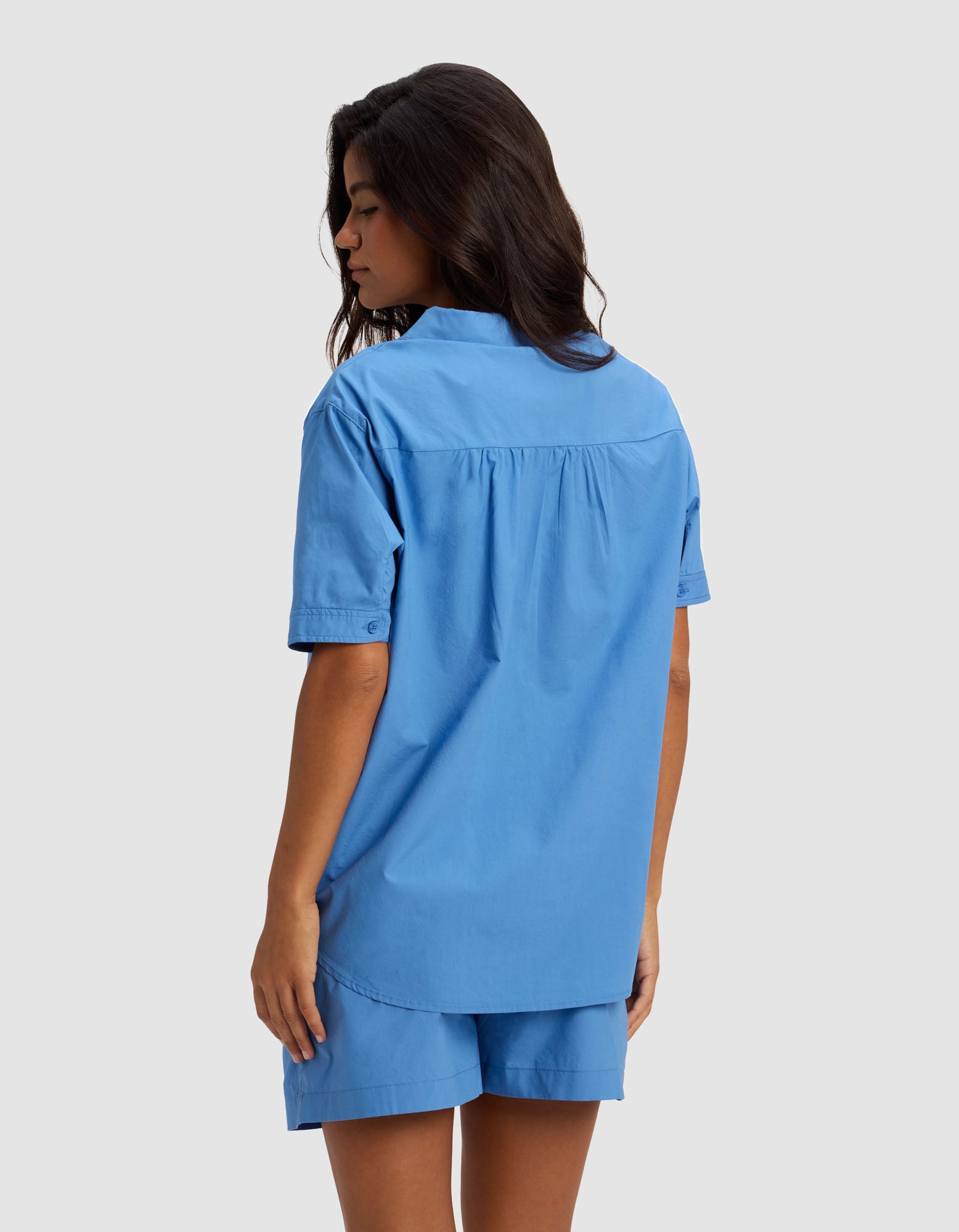 A woman with long dark hair stands sideways, facing away, wearing the Cozy Earth Women's Sutton Short Sleeve Pajama Top in blue and matching shorts against a plain light gray background.|Color:Sailor
