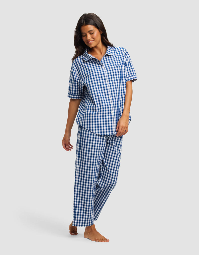 A woman models the Cozy Earth Women's Sutton Short Sleeve & Pant Pajama Set, featuring a blue and white checkered design, standing barefoot against a plain light gray background.