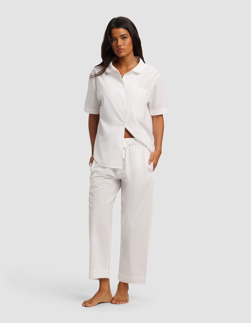 A woman stands barefoot against a plain background, wearing the Cozy Earth Women's Sutton Short Sleeve & Pant Pajama Set in white. The loose-fitting set features a button-up shirt, short sleeves, and matching drawstring pants. Her hands are in her pockets.