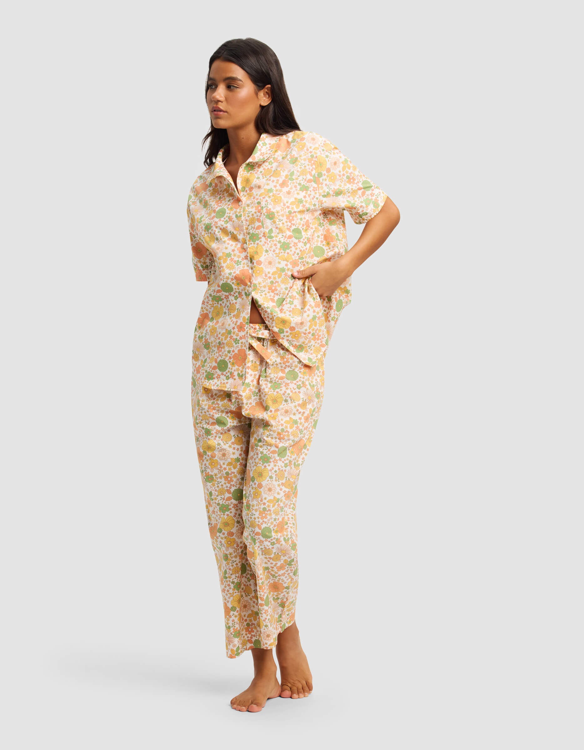 A woman stands barefoot in the Cozy Earth Women's Sutton Short Sleeve & Pant Pajama Set—matching floral pajamas with a pastel short-sleeve button-up top and pants—posing with one hand in her pocket against a plain light background.