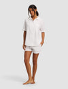 A woman stands barefoot against a light gray background, wearing the Cozy Earth Women's Sutton Short Sleeve Pajama Set—a loose-fitting white button-up shirt with short sleeves and matching white shorts.|Color:White