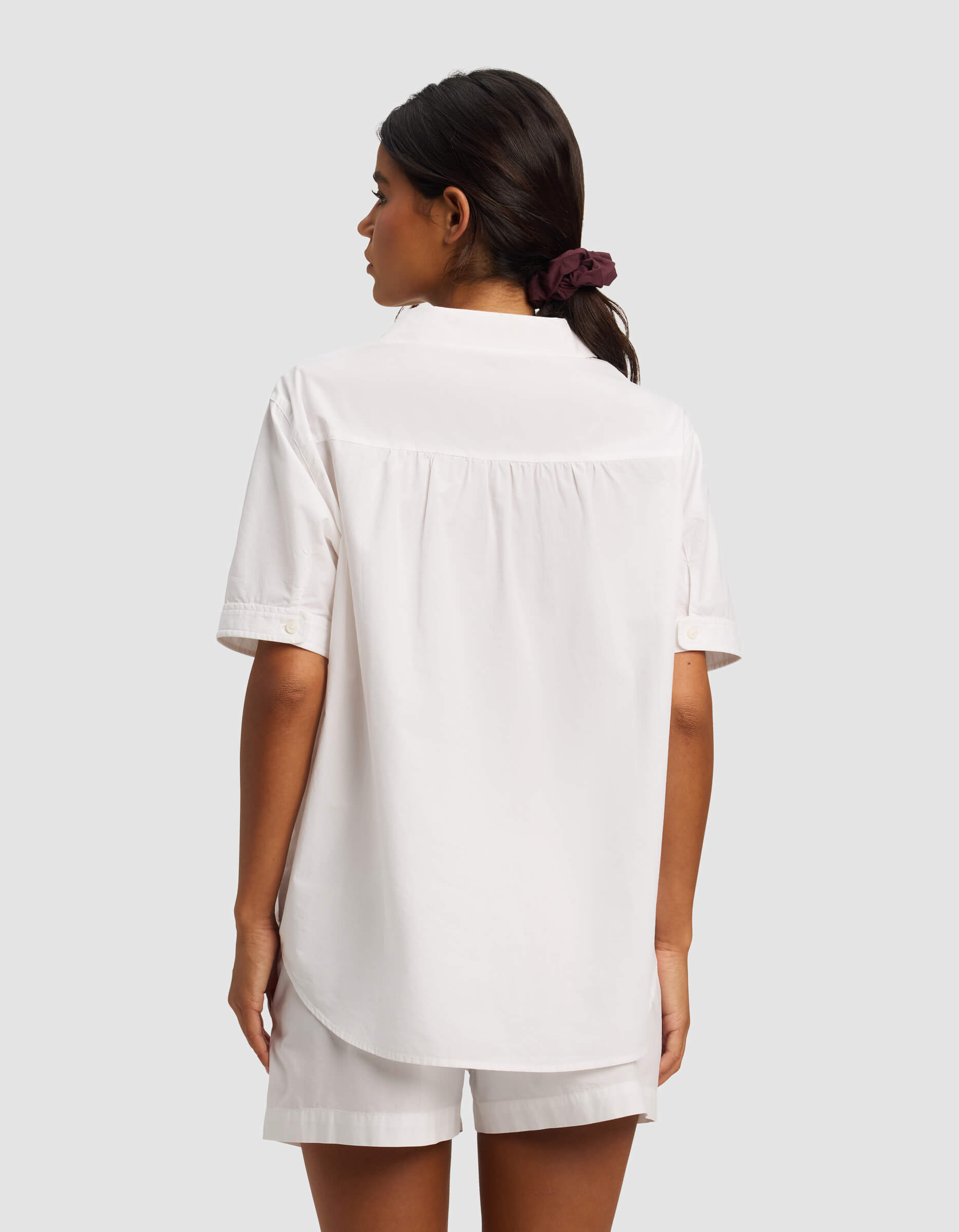 A woman with dark hair in a ponytail, tied with a dark scrunchie, is shown from the back wearing the Cozy Earth Women's Sutton Short Sleeve Pajama Top and matching white shorts against a plain light gray background.