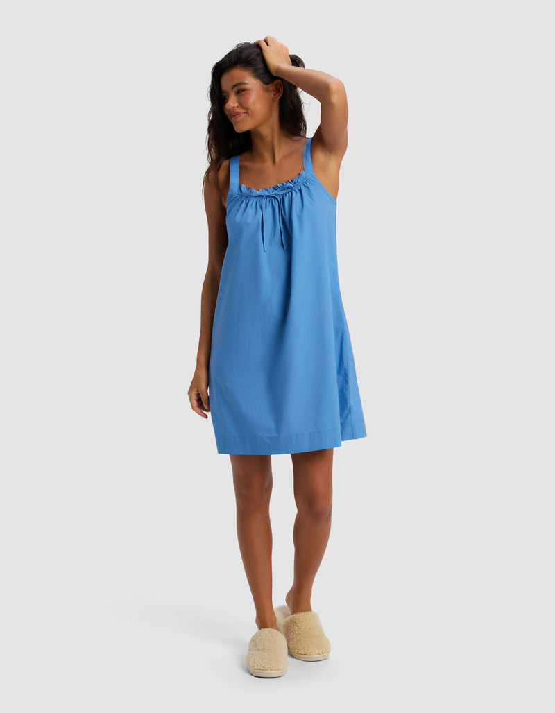 A woman wearing Cozy Earth's Women's Sutton Tank Sleep Dress in blue stands barefoot on a white background, smiling with one hand in her hair and wearing fuzzy beige slippers.