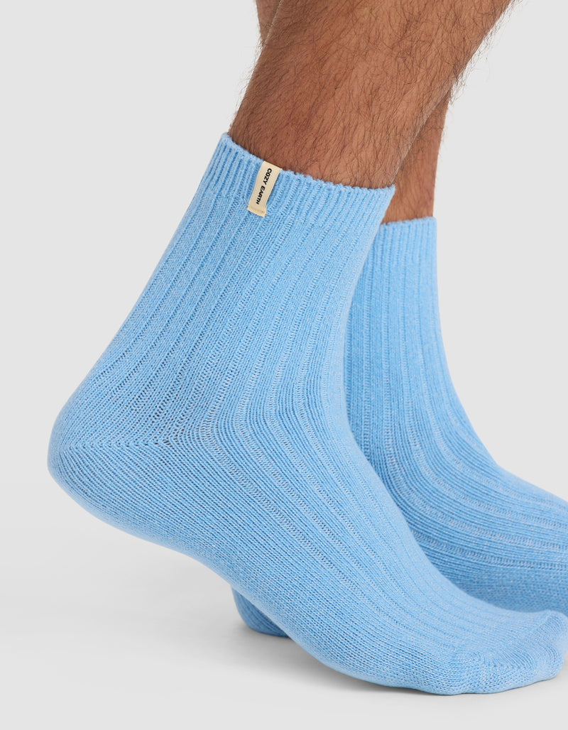 A close-up of a person's lower legs wearing Cozy Earth's Plush Lounge Quarter Sock in light blue with a small beige tag near the top edge, standing on a white background.