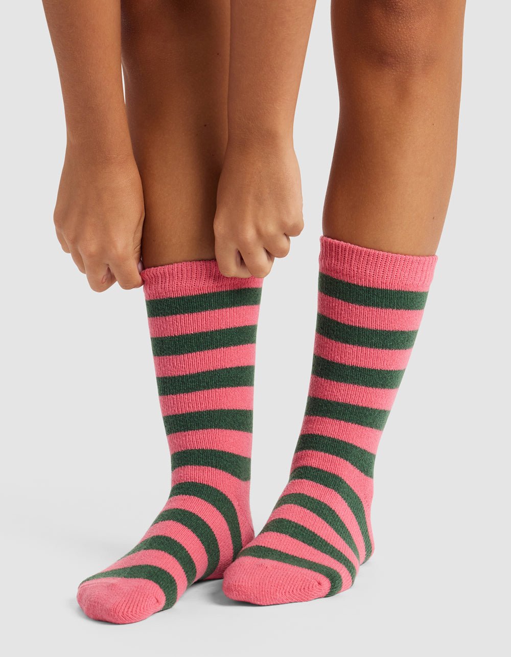 A person stands on a white surface, pulling up The Striped Plush Lounge Sock by Cozy Earth, featuring pink and dark green horizontal stripes. Only their legs and hands are visible.