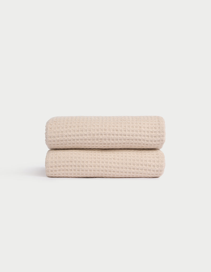 Two Cozy Earth Waffle Hand Towels in light beige are neatly folded and stacked against a plain white background, showcasing their textured pattern.