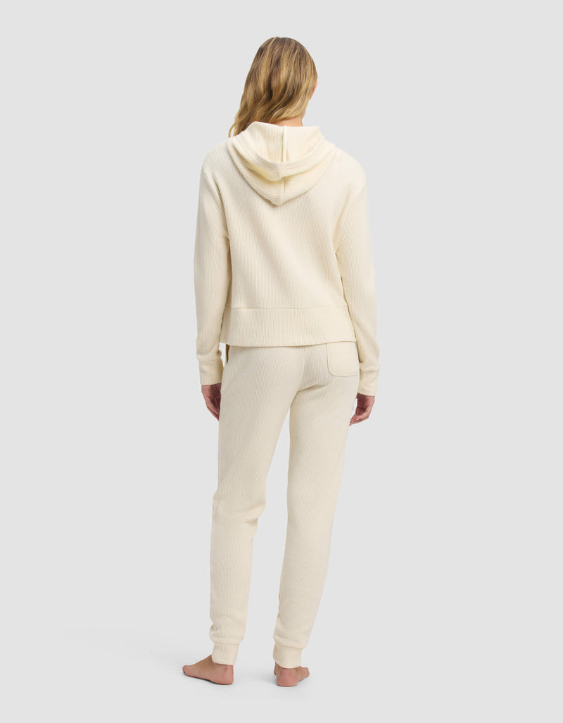 A woman with long blonde hair stands barefoot, facing away, wearing the Cozy Earth Women's Blake Waffle Hoodie & Jogger Set in cream against a plain light gray background.