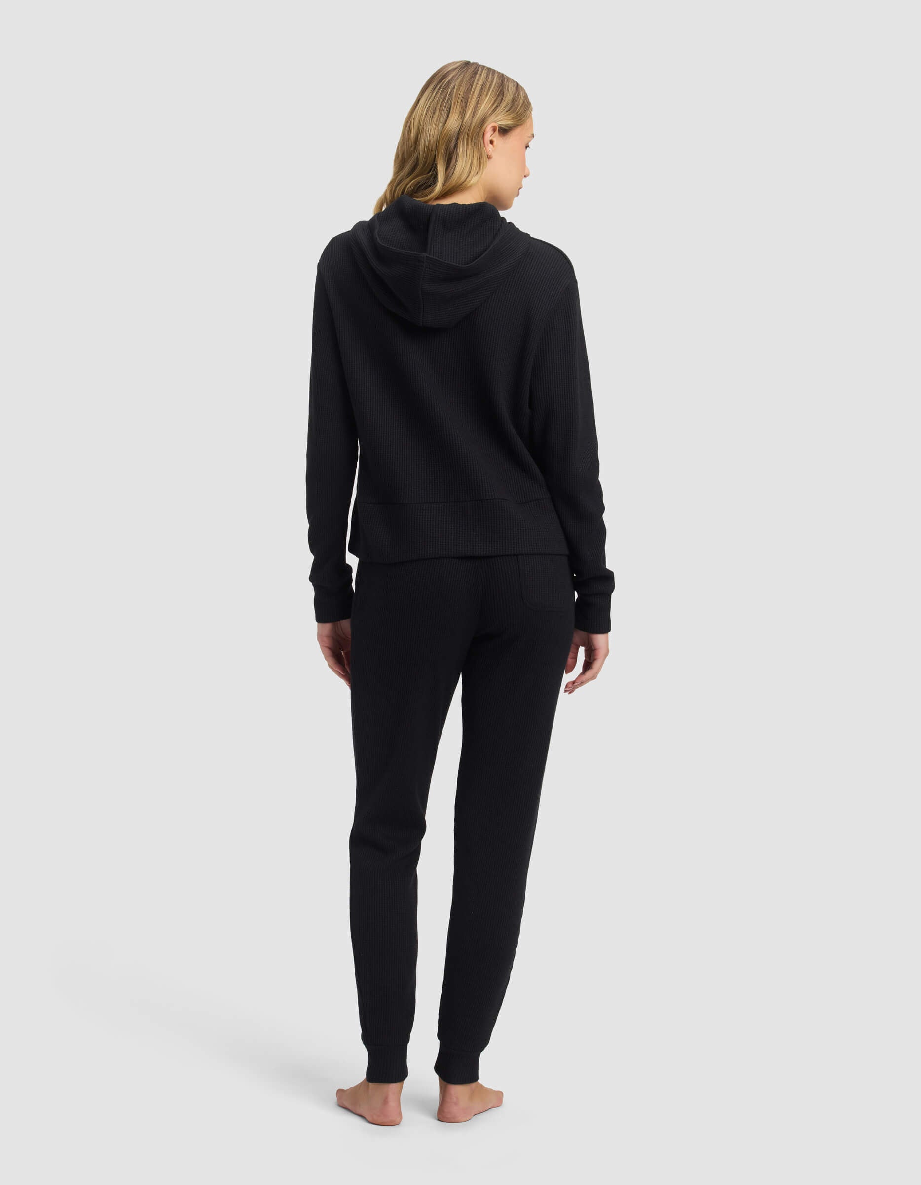 A woman with long blonde hair stands barefoot, facing away from the camera, wearing Cozy Earth's Women's Blake Waffle Hoodie & Jogger Set in black against a plain light gray background.