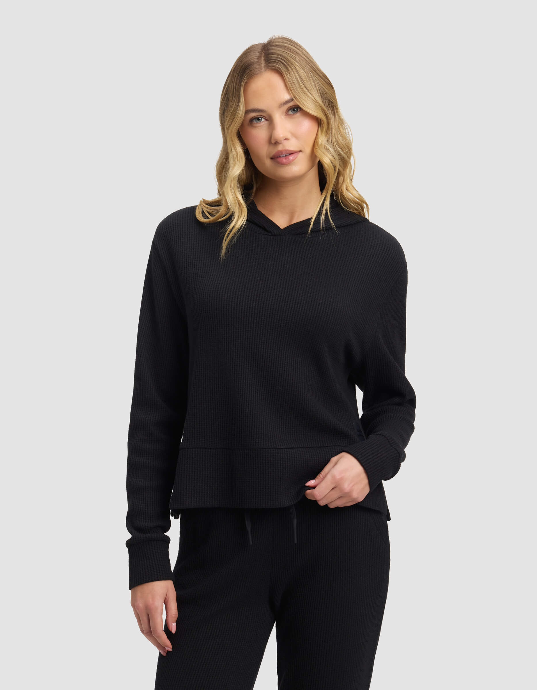 A woman with long, wavy blonde hair wears the Cozy Earth Women's Blake Waffle Hoodie in black, paired with matching black pants, standing against a plain light gray background.|Color:Jet Black