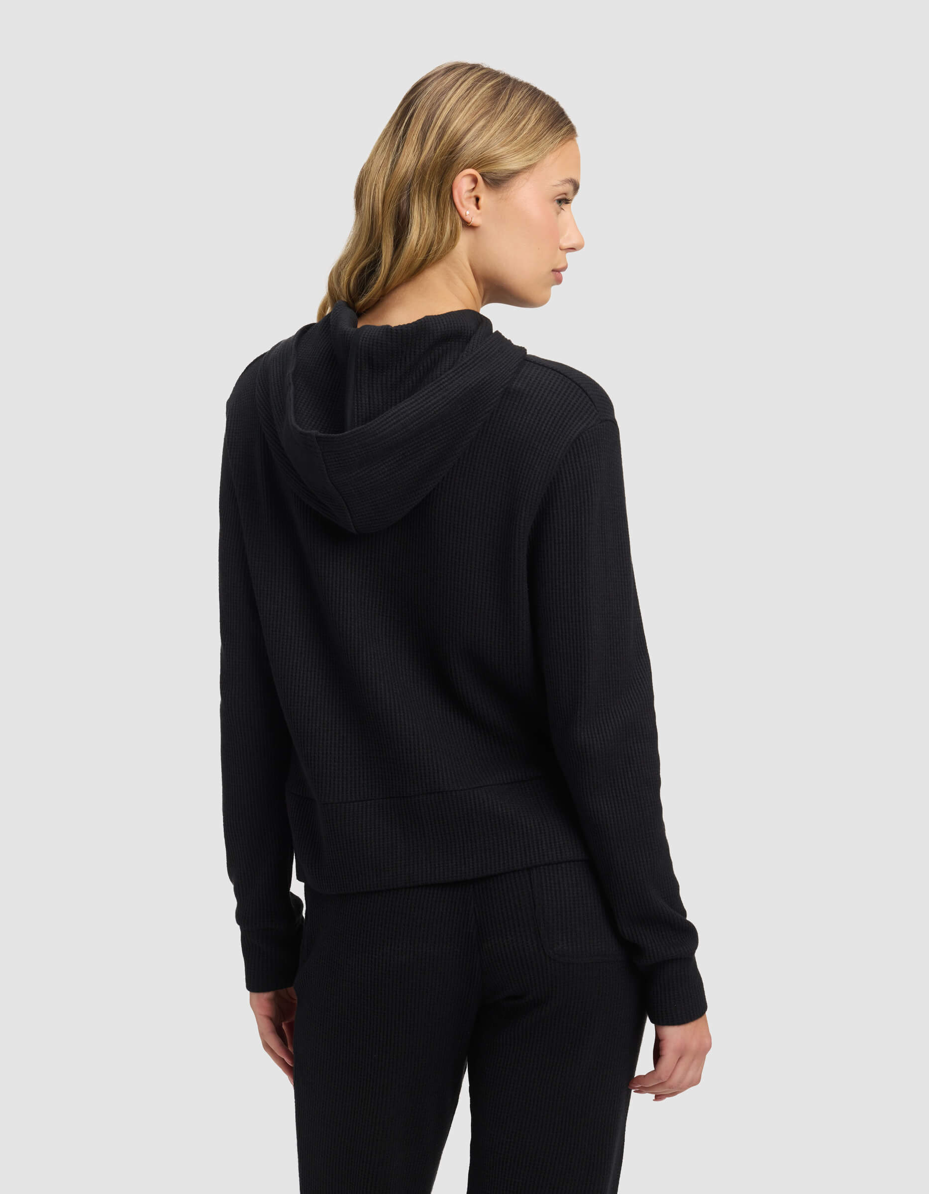 A woman with blonde, wavy hair is seen from the back wearing the Cozy Earth Women's Blake Waffle Hoodie and matching pants, standing against a plain light gray background.