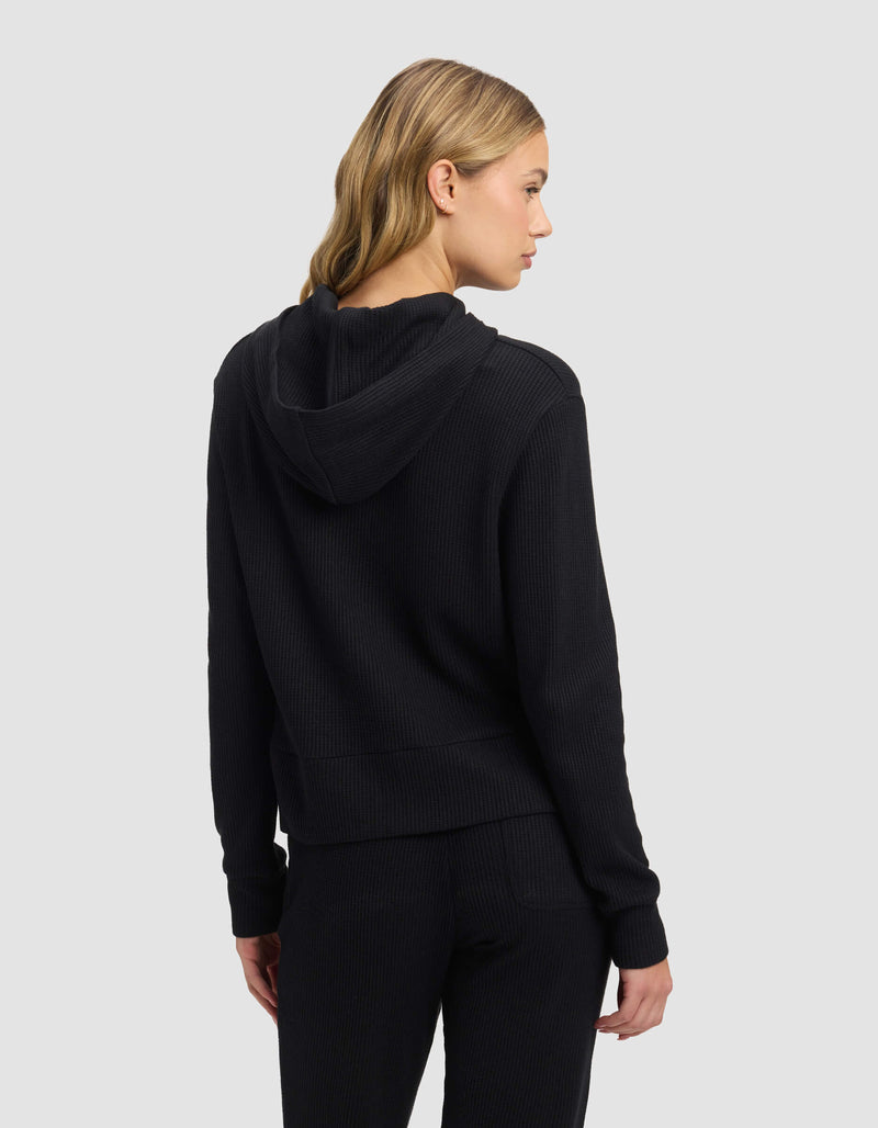 A woman with blonde, wavy hair is seen from the back wearing the Cozy Earth Women's Blake Waffle Hoodie and matching pants, standing against a plain light gray background.