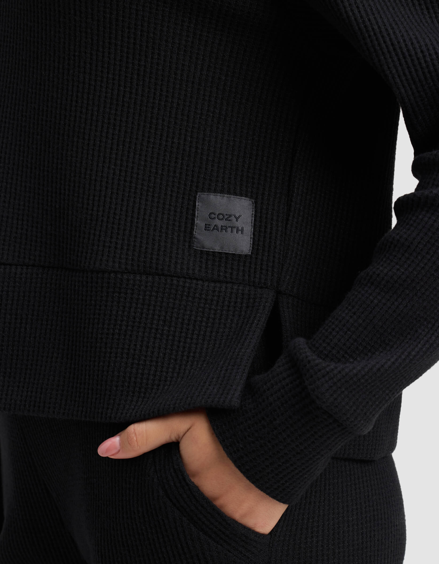 A woman wears the Cozy Earth Women's Blake Waffle Hoodie in black, hand in pocket; a gray patch on the shirt displays the brand name.