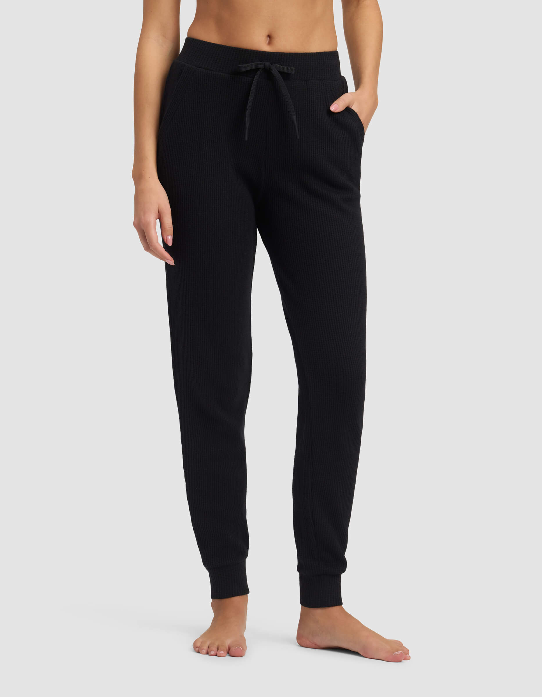 A person stands barefoot against a light gray background, modeling the lower torso and legs in Cozy Earth's Women's Blake Waffle Jogger, featuring a ribbed texture, drawstring waist, and pockets.|Color:Jet Black