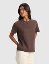 A woman with long dark hair wears Cozy Earth's Women's All Day Tee in brown and off-white pants, standing against a light gray background with her hands behind her back, looking to her left.|Color:Cacao