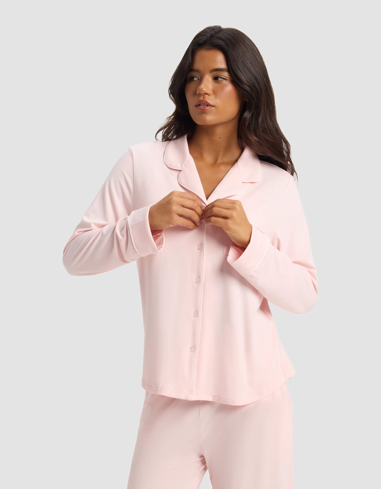 A woman with long dark hair wears the Cozy Earth Women's Bamboo Stretch-Knit Long Sleeve Pajama Set in light pink, fastening a button on her top as she stands against a plain light gray background, looking slightly to her left.
