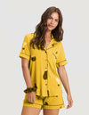 A woman with wavy brown hair wears the Cozy Earth Women's Bamboo Stretch-Knit Short Sleeve Pajama Set (Veggie), featuring a button-up top, matching shorts, and a coordinating scrunchie. She looks relaxed. |Color:Veggies in Mango