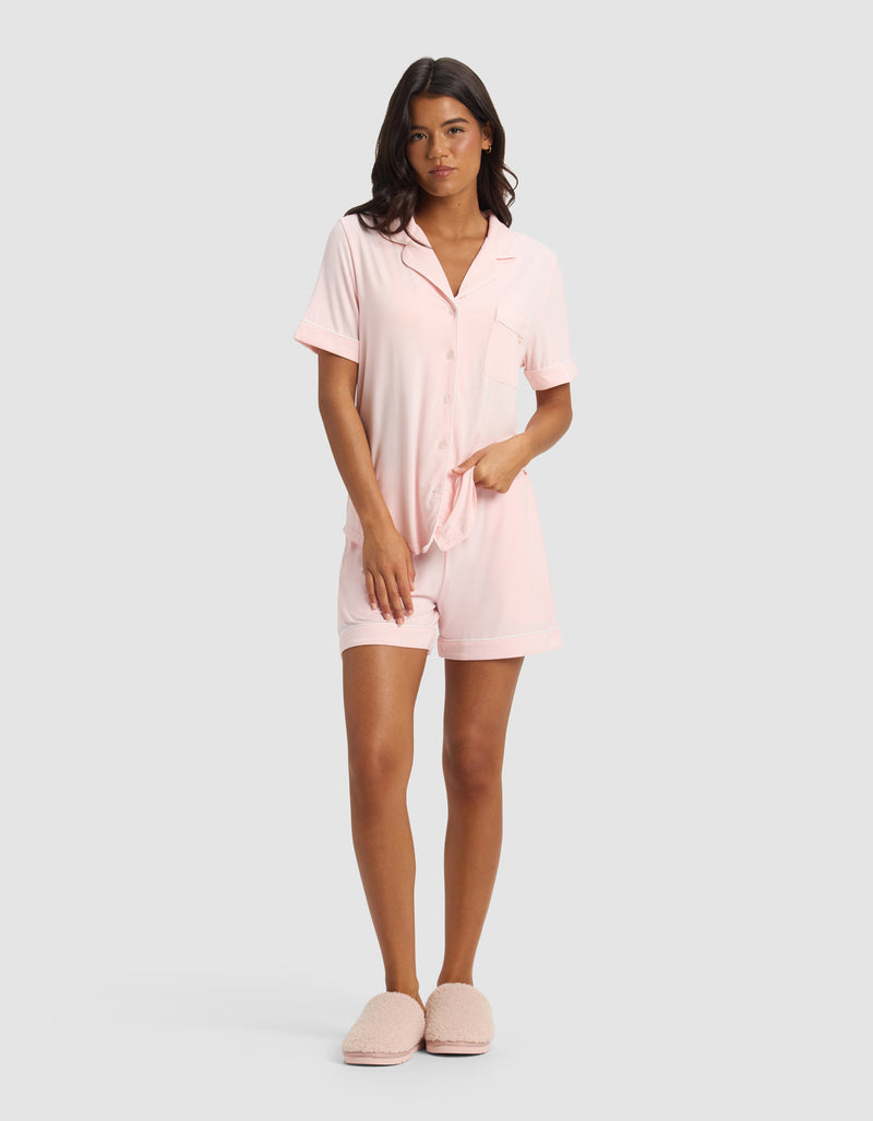 A woman faces forward wearing the Cozy Earth Women's Bamboo Stretch-Knit Short Sleeve Pajama Set in light pink with matching shorts and slippers, set against a plain light gray background.