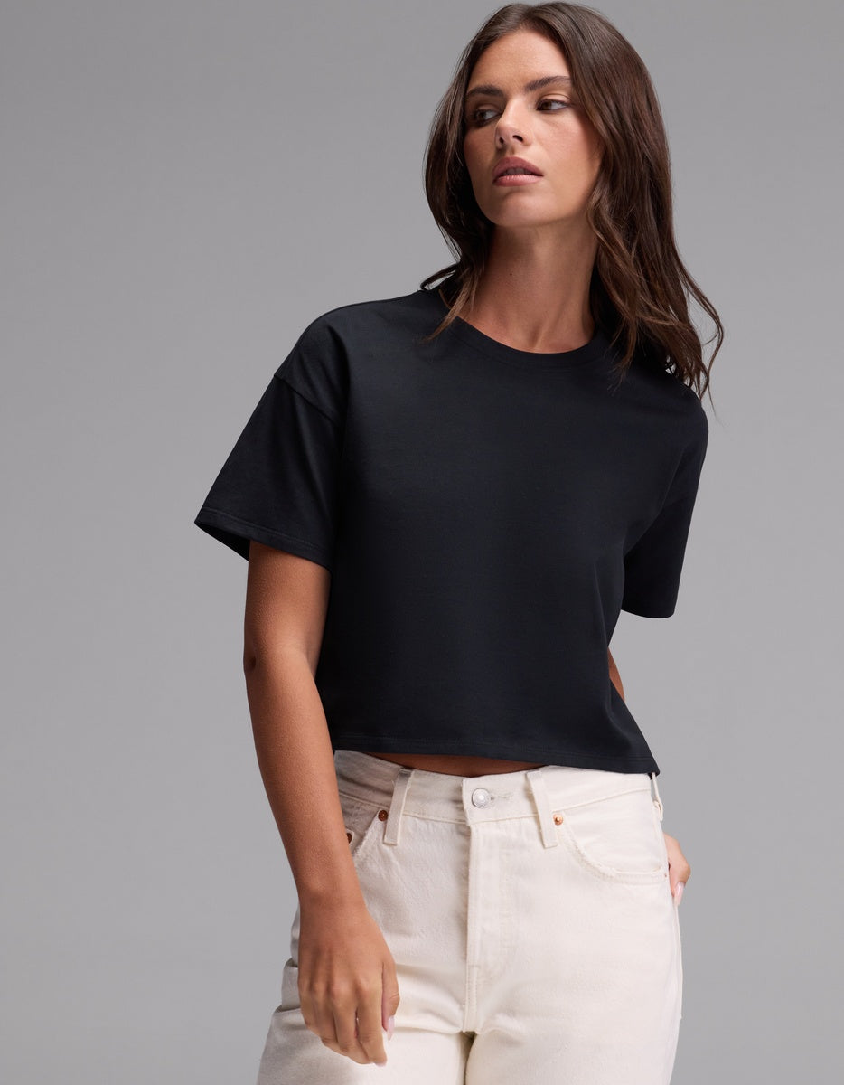A woman stands against a plain gray background, wearing the Cozy Earth Women’s All Day Cropped Tee in Jet Black paired with high-waisted white jeans, with one hand casually placed in her pocket.|Color:Jet Black
