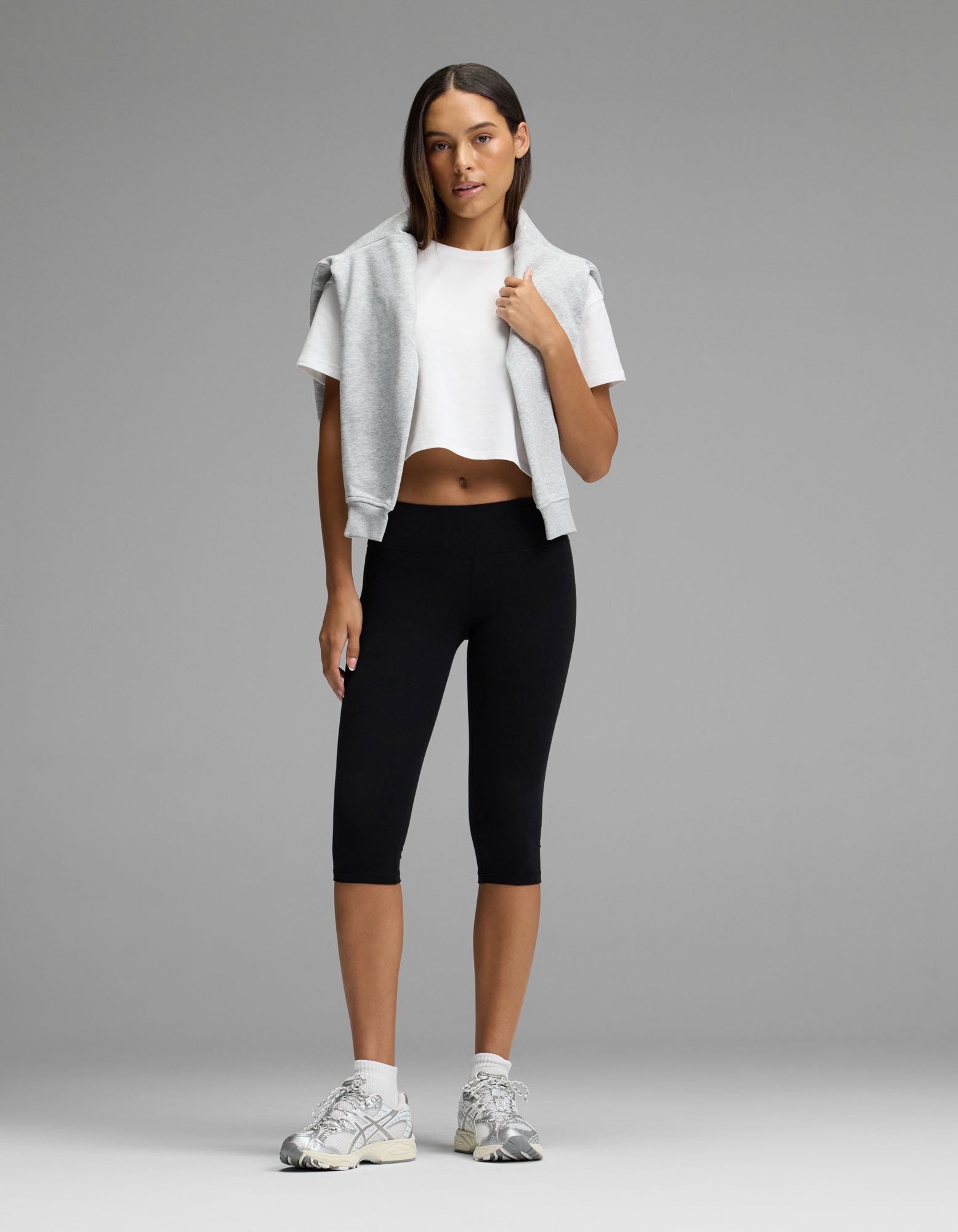 A woman stands against a gray background, wearing the Women’s All Day Cropped Tee by Cozy Earth, paired with black leggings, white sneakers, and a gray sweatshirt draped over her shoulders. Her straight hair and relaxed, confident pose complete the look.